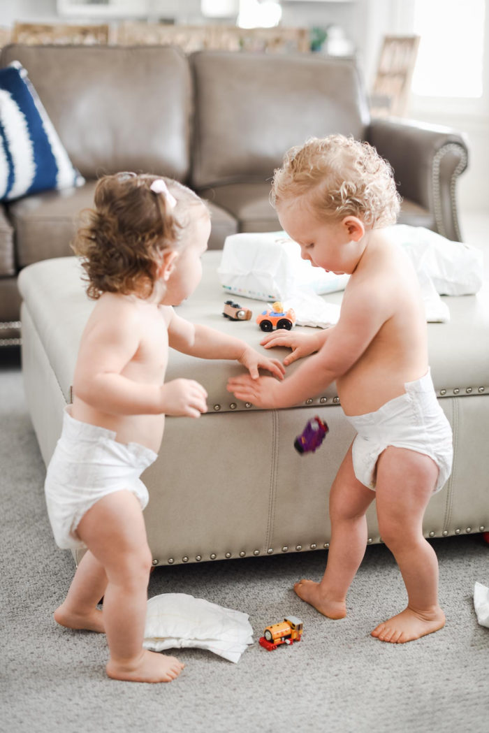 Walmart Baby Registry Must-Have by popular Utah life and style blog, A Slice of Style: image of a fraternal boy and girl twins wearing only diapers and playing with cars and trains on a large light grey ottoman that has some Pamper's Pure Protection diapers from Walmart on it.