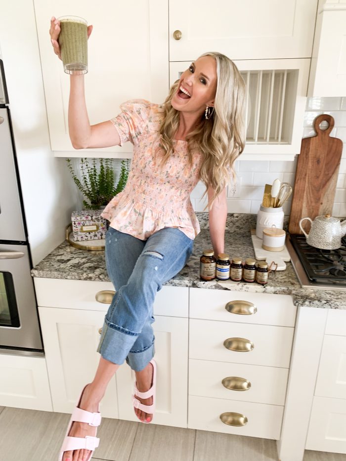 Solgar Vitamins Review + Green Smoothie Recipe by popular Utah life and style blog, A Slice of Style: image of a woman sitting on her kitchen counter next to various bottle of Solgar vitamins and holding a green smoothie in a clear tumbler cup.